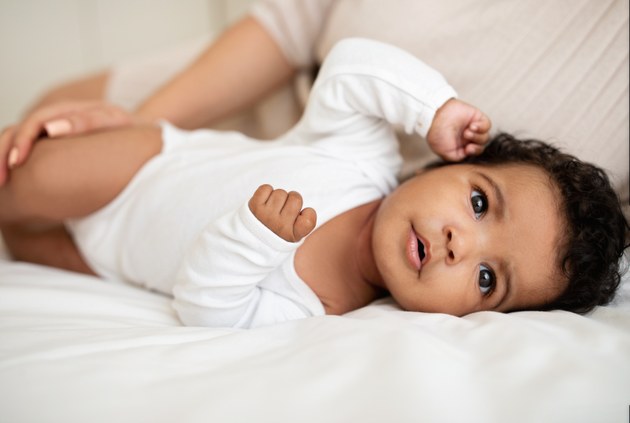 Baby mit dunkler Hautfarbe und schwarzen Haaren liegt seitlich auf einem weißen Tuch und schaut zum Betrachter. Auf seinem Bein eine Hand mit langen Fingernägeln.
