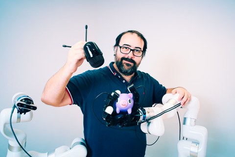 Fotoaufnahme von Roberto Calandra, der neben einem Roboterarm steht. Dieser hat ein lilafarbenes Spielzeugschweinchen in der Hand. Roberto Calandra hält ein Mikrofon in einer Hand.
