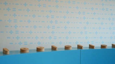Blauer Sockel mit Holzzylindern in verschiednen Größen vor heller Wand. 