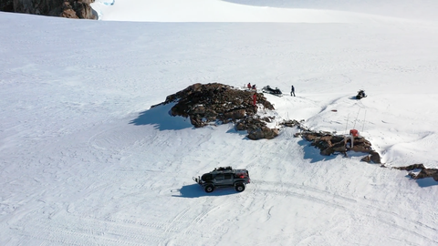 Personen arbeiten auf einem Felsen, der von Schnee und Eis umgeben ist. Zu sehen sind auch Schneemobile und ein Auto.