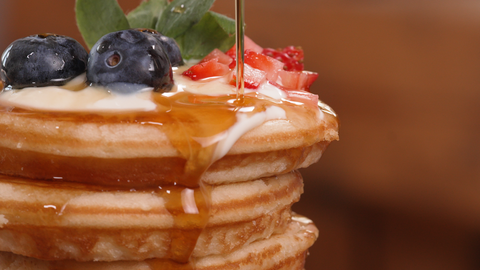 Foto von einem Stapel Pancakes, der mit frischen Blaubeeren und gehackten Erdbeeren garniert ist. Über die Pancakes wird gerade Sirup gegossen, der über die Seiten hinunterläuft.