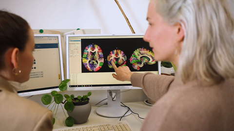 Foto von zwei Frauen an einem Schribtisch, die sich unterhalten. Auf den Bildschirmen vor ihnen sind bunte CT-Aufnahmen zu sehen.