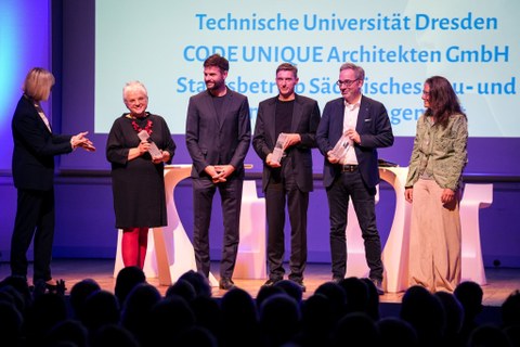 Foto einer Preisübergabe: auf einer Bühne stehen 5 lächelnde Personen. Links steht eine ihnen zugewandte Person, die Hände nach vorn gestreckt.