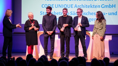 Foto einer Preisübergabe: auf einer Bühne stehen 5 lächelnde Personen. Links steht eine ihnen zugewandte Person, die Hände nach vorn gestreckt.