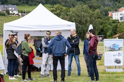 Foto einer Gruppe von Menschen, die einander zugewandt vor einem Pavillon mit weißem Stoffdach stehen.