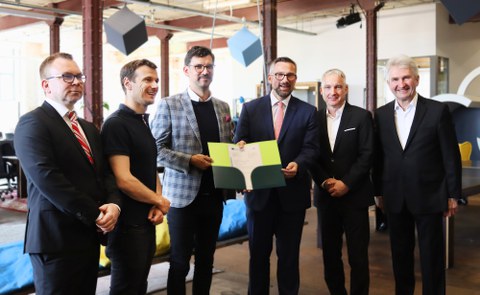 Gruppenbild mit 6 Herren nebeneinander im Anzug. Die zwei Herren in der MIttel halten gemeinsam eine Urkunde in einer Mappe in den Händen. Alle Herren schauen freundlich.