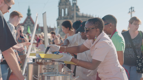 Fotoaufnahme von Menschen, die bei einem Fest im Freien vor der Kulisse der Dresdner Alstadt Essen aus großen Kochtöpfen servieren.