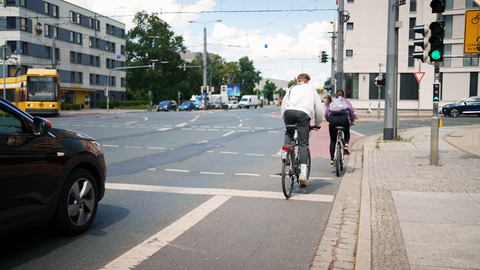 Foto von zwei Radfahrer:innen, die auf dem Radweg eine Kreuzung überqueren. Neben ihnen ist steht ein Auto. Auf der Kreuzung biegt eine Straßenbahn ab.