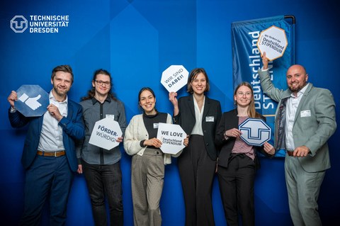 Vor einer blauen Wand mit dem weißen TUD-Logo oben links stehen sechs lachende Personen. Diese halten jeweils eine eckige Karte mit verschiedenen Symbolen und Wörtern, die im Zusammenhang mit der TU Dresden und den Deutschlandstipendium stehen, in der Hand.