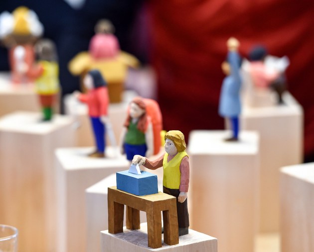 Verschiedene Holzfiguren zur Darstellung der Gesellschaft. Im Vordergrund eine Frau an der Wahlurne.