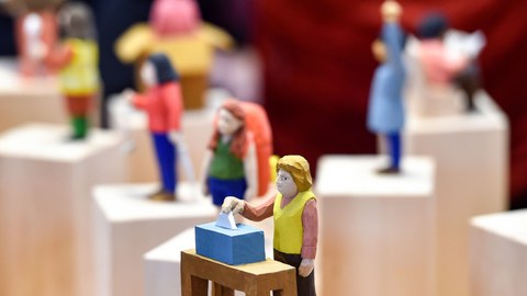 Verschiedene Holzfiguren zur Darstellung der Gesellschaft. Im Vordergrund eine Frau an der Wahlurne.