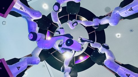 Blick von unten auf 6 weiß-schwarze Roboterarme mit runden Konturen, die sich, von außen kommend, in der Mitte treffen. Sie sind violett angestrahlt. Im Hintergrund eine weiße Zimmerdecke, in deren Mitte ein schwarzer Ring, der in 6 Segmente geteilt ist.