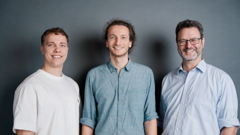 Porträtfoto von drei jungen Männern, die nebeneinander stehen und in die Kamera blicken.