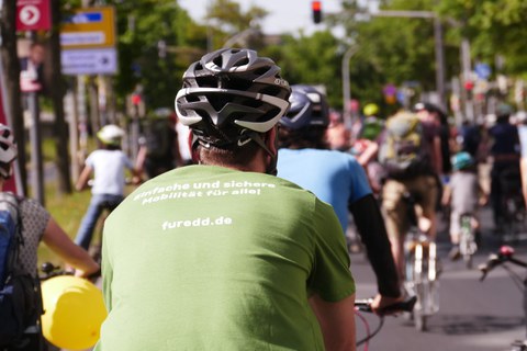 Foto von Kopf und Rücken eines Fahrradfahrers. Er hat einen grau-weißen Fahrradhelm und ein hellgründes T-Shirt mit der weißen Aufschrift: Einfache und sichere, darunter: Mobilität für alle! Darunter: furedde.de. Vor ihm sind verschwommen andere Fahrradfahrer von hinten zu sehen.