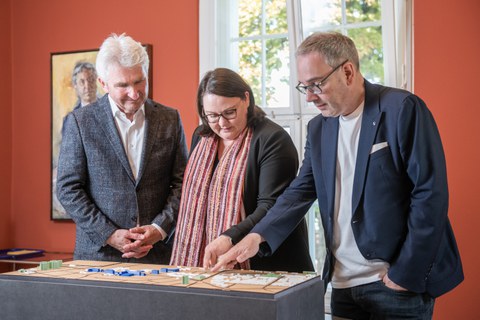 Foto zweier Herren rechts und links und einer Dame in der Mitte, die auf ein vor ihnen stehendes, dreidimensionales Modell einer Stadt schauen und darauf zeigen.