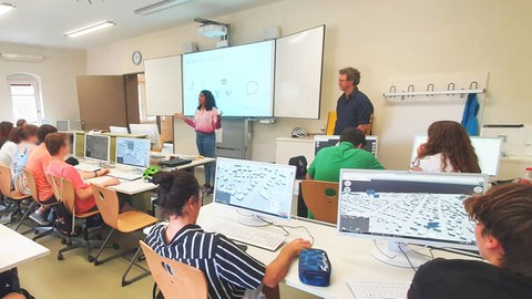 Gymnasiasten im Unterricht, vor der Tafel steht eine Lehrerin, daneben ein Mann.