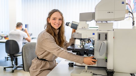 Fotoaufnahme einer Frau, die an einem Mikroskop sitzt und in die Kamera lächelt.