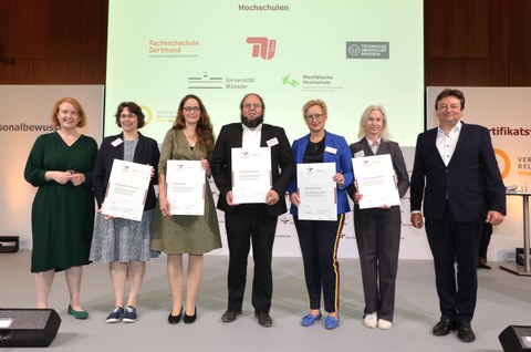 Sieben Menschen stehen nebeneinander und halten Zettel vor ihrem Bauch