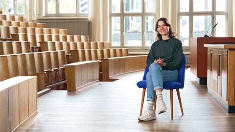 Foto einer lächelnden Frau, die in einem Hörsaal auf einem blauen Stuhl sitzt.