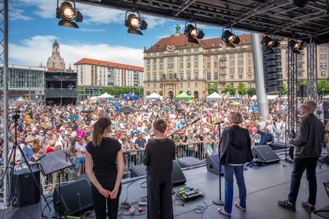 Foto von TUD-Rektorin Ursula Staudinger in drei weiteren Personen, die auf einer Bühne auf dem Dresdner Altmarkt stehen und auf eine Menschenmenge blicken. Im Hintergrund ist der Dresdner Kulturpalast sowie die Frauenkirche abgebildet.