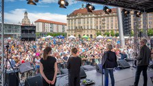 Foto von TUD-Rektorin Ursula Staudinger in drei weiteren Personen, die auf einer Bühne auf dem Dresdner Altmarkt stehen und auf eine Menschenmenge blicken. Im Hintergrund ist der Dresdner Kulturpalast sowie die Frauenkirche abgebildet.
