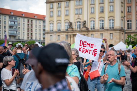 Fotoaufnahme von Veranstaltungsteilnehmenden auf dem Dresdner Altmarkt. Eine Frau hält ein Transparent ins Bild mit dem Schriftzug Demokratie und einem Herz.