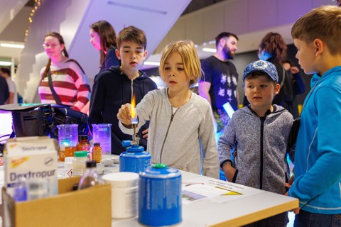 Foto von einem Jungen, der etwas in einen Bunsenbrenner hält. Weitere Personen stehen um ihn herum und beobachten das Geschehen.
