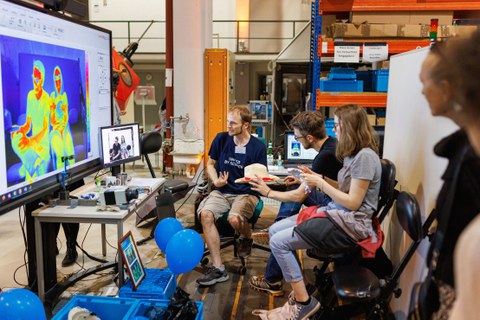 Foto von zwei Personen, die auf Stühlen sitzen. Vor ihnen befindet sich ein Monitor, der sie im Wärmebild zeigt.