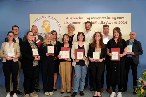 Foto von 14 Personen, die in drei Reihen vor einer blauen Wand stehen. Darauf steht auf rechteckigem, hellgelben Untergrund rechts der Schriftzug: Auszeichnungsveranstaltung zum, darunter 29. Comenius EduMedia Award-2024. Auf der linken Seite das runde Logo der Comenius Gesellschaft