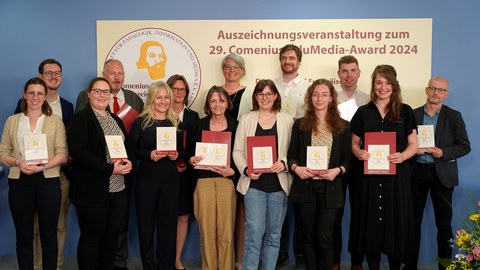 Foto von 14 Personen, die in drei Reihen vor einer blauen Wand stehen. Darauf steht auf rechteckigem, hellgelben Untergrund rechts der Schriftzug: Auszeichnungsveranstaltung zum, darunter 29. Comenius EduMedia Award-2024. Auf der linken Seite das runde Logo der Comenius Gesellschaft