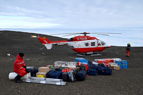 Auf einem felsen steht ein rot-weißer Hubschrauber, davor sind viele Gepäckstücke aufgebaut.