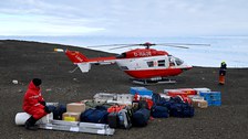 Auf einem felsen steht ein rot-weißer Hubschrauber, davor sind viele Gepäckstücke aufgebaut.