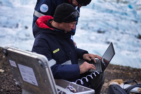 Ein Mann sitzt mit Laptop auf eijnem Felsen im Schnee. Deneben steht ein Koffer mit Tecnik.