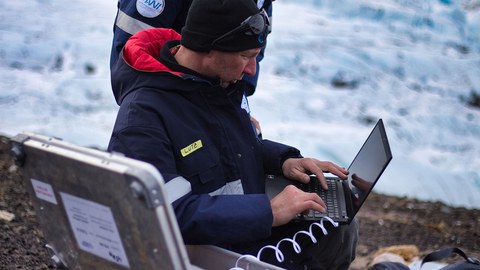 Ein Mann sitzt mit Laptop auf eijnem Felsen im Schnee. Deneben steht ein Koffer mit Tecnik.