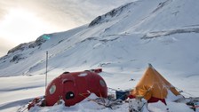 Ein gelbes Zelt und eine rote Wohnkapsel stehen im Schnee vor einem Berg.