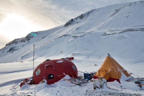 Ein gelbes Zelt und eine rote Wohnkapsel stehen im Schnee vor einem Berg.