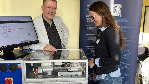 Junge Frau blickt interessiert auf einen Messestand. Sort steht auf einem Tisch der kastenförmige, gläserne Demonstrator LOTTA. Hinter dem Tisch steht ein Referent am Messestand..ein 