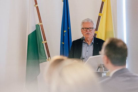 Foto eines Herrn mit weißen Haaren und Schnauzbart und Brille vor drei Flaggen stehend. v.l. weiß-grün, blau, gelb. Im Vordergrund sind die verschwommenen Hinterköpfe zweier Personen zu sehen.