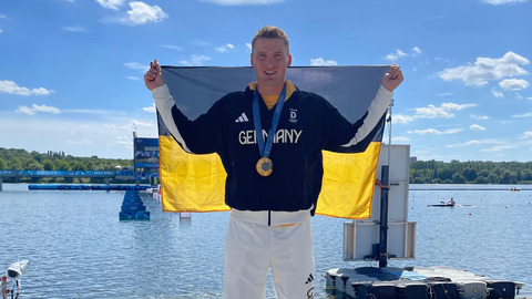 Fotoaufnahme von Tom Liebscher-Lucz, der in einem Trainingsanzug des deutschen Olympia-Teams vor der Kanu-Rennstrecke steht. Er hat eine Gold-Medaille um den Hals hängen und trägt hinter seinem Rücken eine schwarz-gelbe Fahne.