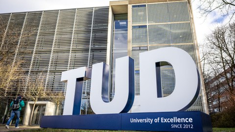 Fotografie von einer großen Skulptur aus drei großen weißen 3D-Buchstaben „TUD“ auf blauem Sockel auf einer Wiese im Hintergrund das Hörsaalzentrum.