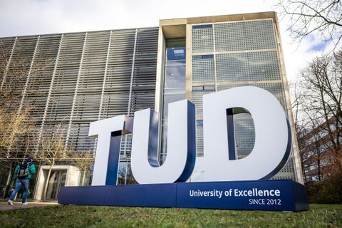 Photograph of a large sculpture of three large white 3D letters "TUD" on a blue pedestal. The sculpture is located on a lawn with the Auditorium Center in the background.