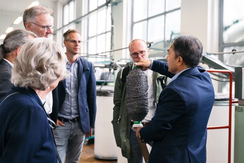 Foto von mehreren Personen, die an einer Führung in den Laboren des ITM teilnehmen. Eine Person hat eine Art Spindel in denm Händen und zeigt das aufgewickelte Material den umstehenen Personen.