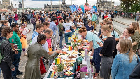 Foto von zahlreichen Personen, die sich entlang einer langen Essenstafel auf der Augustusbrücke in Dresden befinden. Im Hintergrund sind Gebäude der Dresdner Altstadt zu erkennen.