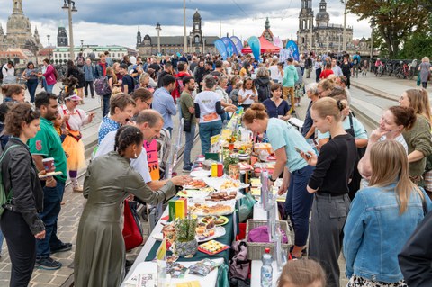 Foto von zahlreichen Personen, die sich entlang einer langen Essenstafel auf der Augustusbrücke in Dresden befinden. Im Hintergrund sind Gebäude der Dresdner Altstadt zu erkennen.