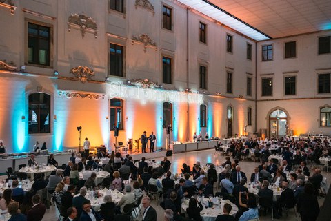 Foto einer Abendveranstaltung im Dresdner Albertinum. In der Bildmitte ist eine Bühne erkennbar, auf der zwei Personen stehen. Im Raum befinden sich viele runde Tische, an denen Teilnehmende der Veranstaltung sitzen. 