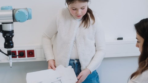 Ein junges Mädchen hält Papiere mit Skizzen in der Hand, die von einem Roboterarm angefertigt wurden.