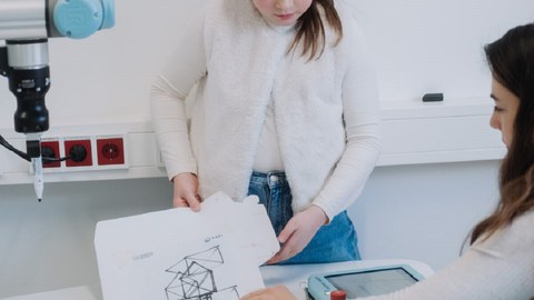 Ein junges Mädchen hält Papiere mit Skizzen in der Hand, die von einem Roboterarm angefertigt wurden.