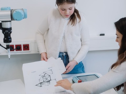 Ein junges Mädchen hält Papiere mit Skizzen in der Hand, die von einem Roboterarm angefertigt wurden.
