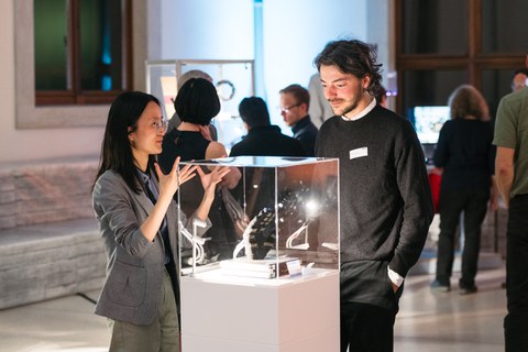 Foto von einer Frau und einem Mann, die an einer Vitrine stehen, die einen Handschuh ausstellt. Die beiden Personen befinden sich in einem Gespräch.