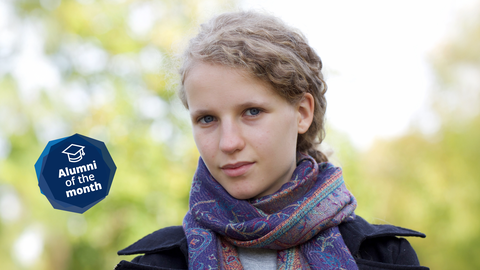 Porträtfoto von Lilith Diringer im Freien. Im Hintergrund ist unscharf das Laub von Bäumen zu erkennen. Das Bild enthält eine blaue Grafik mit dem Text "Alumni of the Month".
